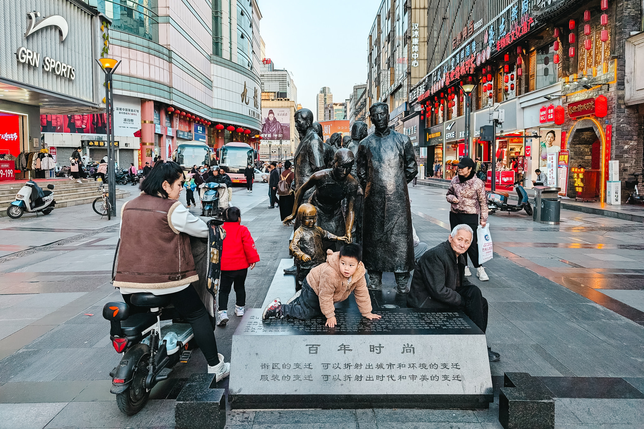 Bronze statue group on urban street with people and scooters