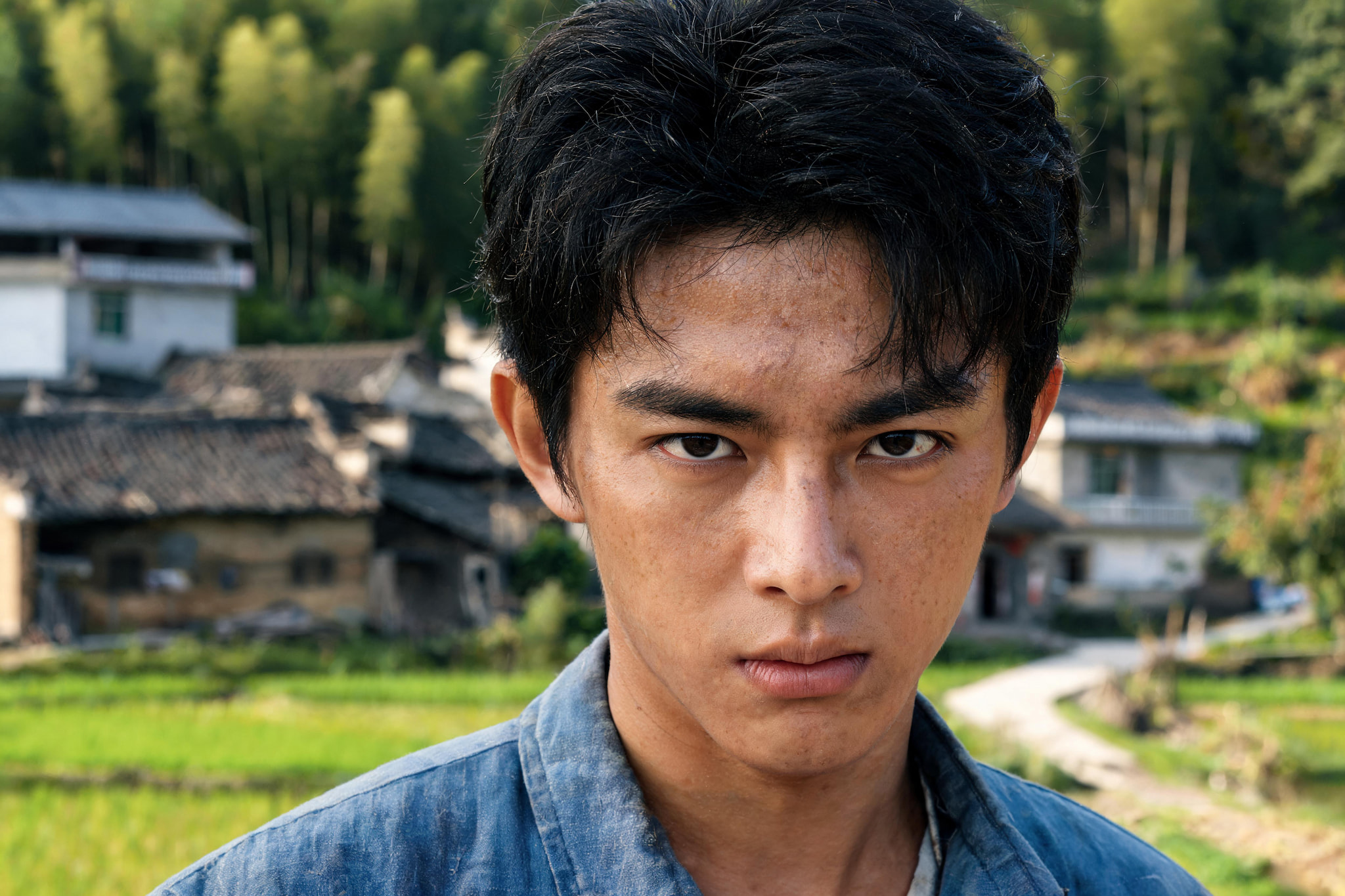 A close-up portrait of a young man with a serious expression in a rural setting.