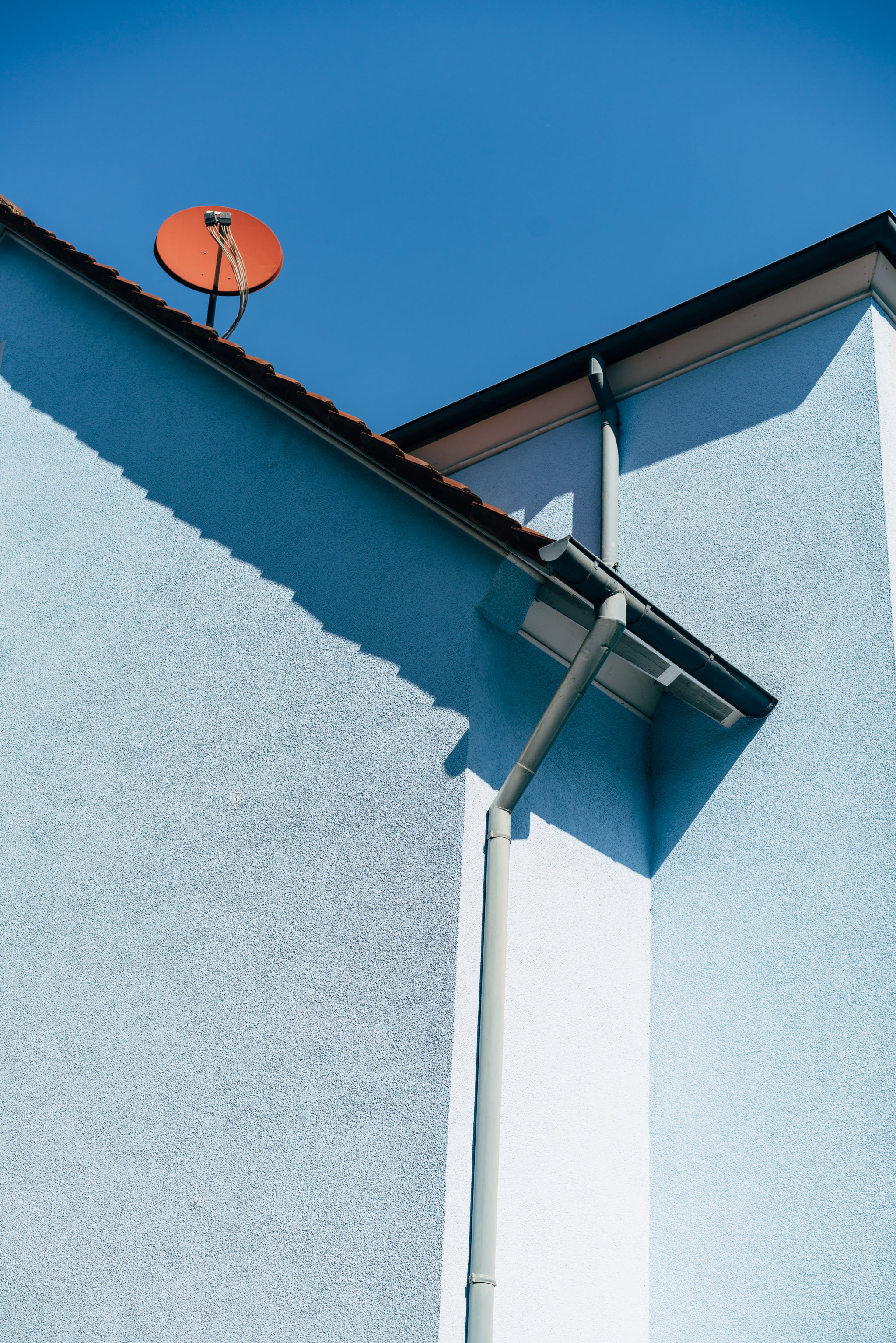 A low-angle view of a blue building exterior with a red satellite dish and drainage pipe against a clear sky.