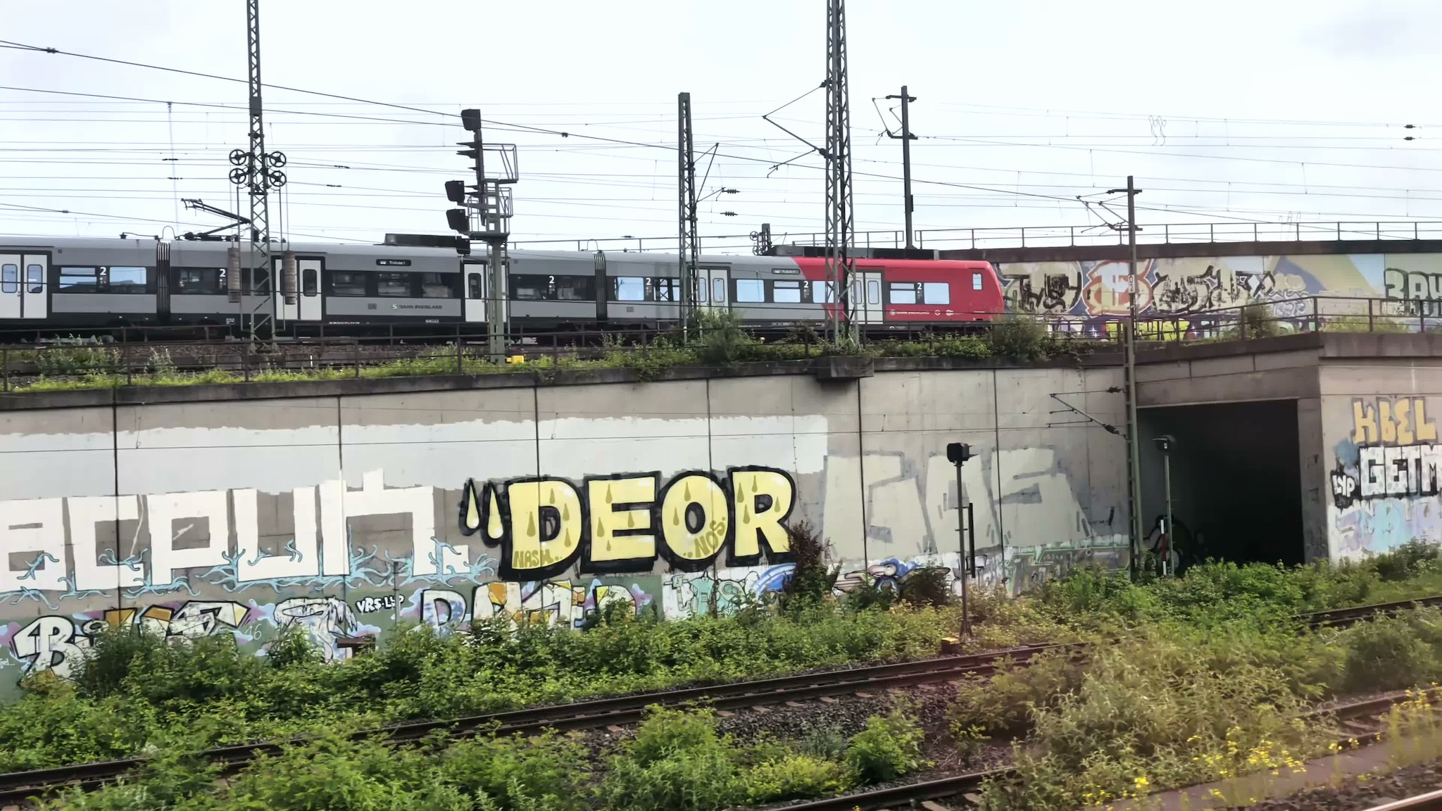 A red and grey commuter train passing over a concrete wall covered in extensive graffiti near train tracks.