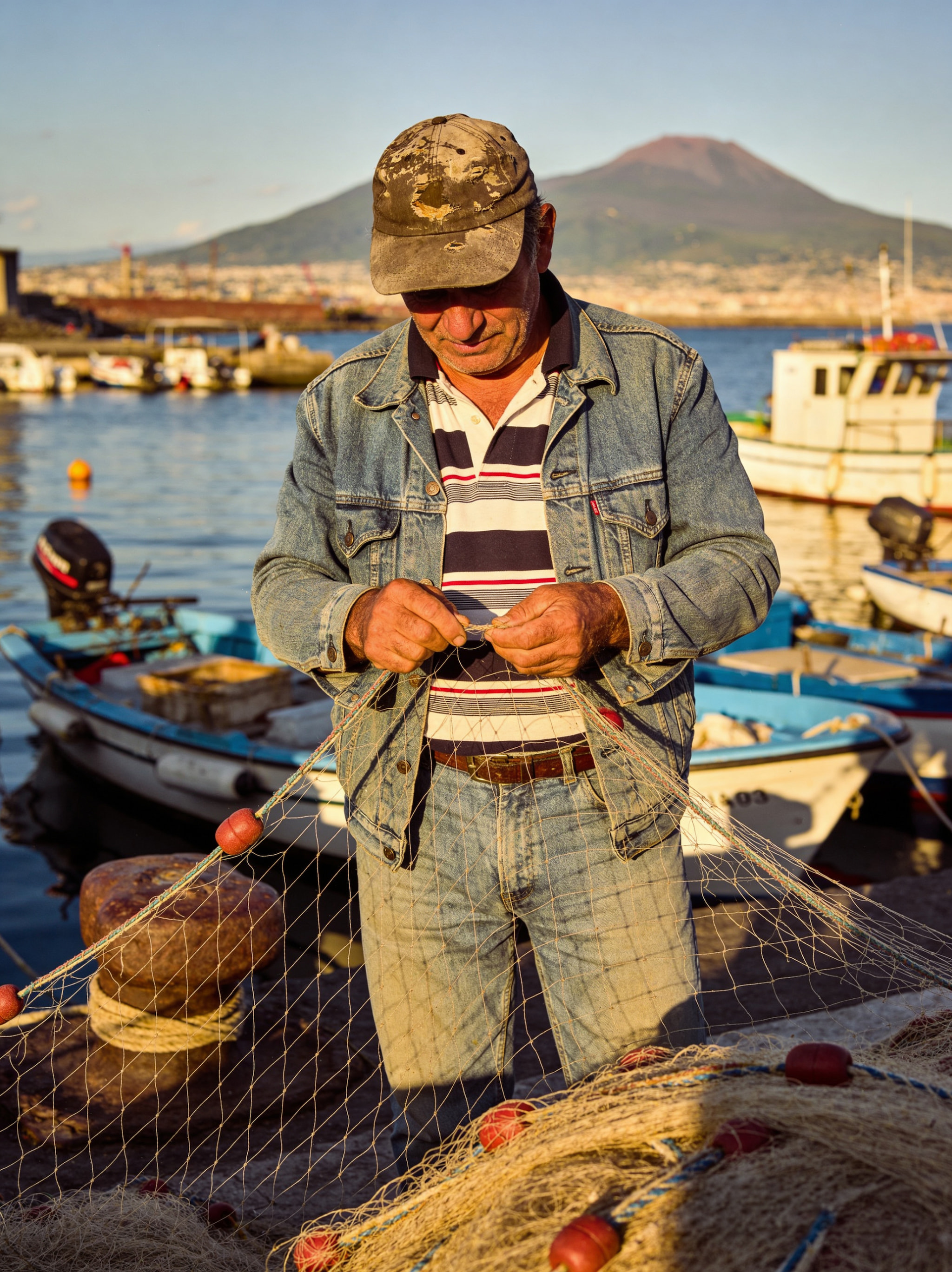 An elderly fisherman mending a net by the harbor with Mount Vesuvius in the background.