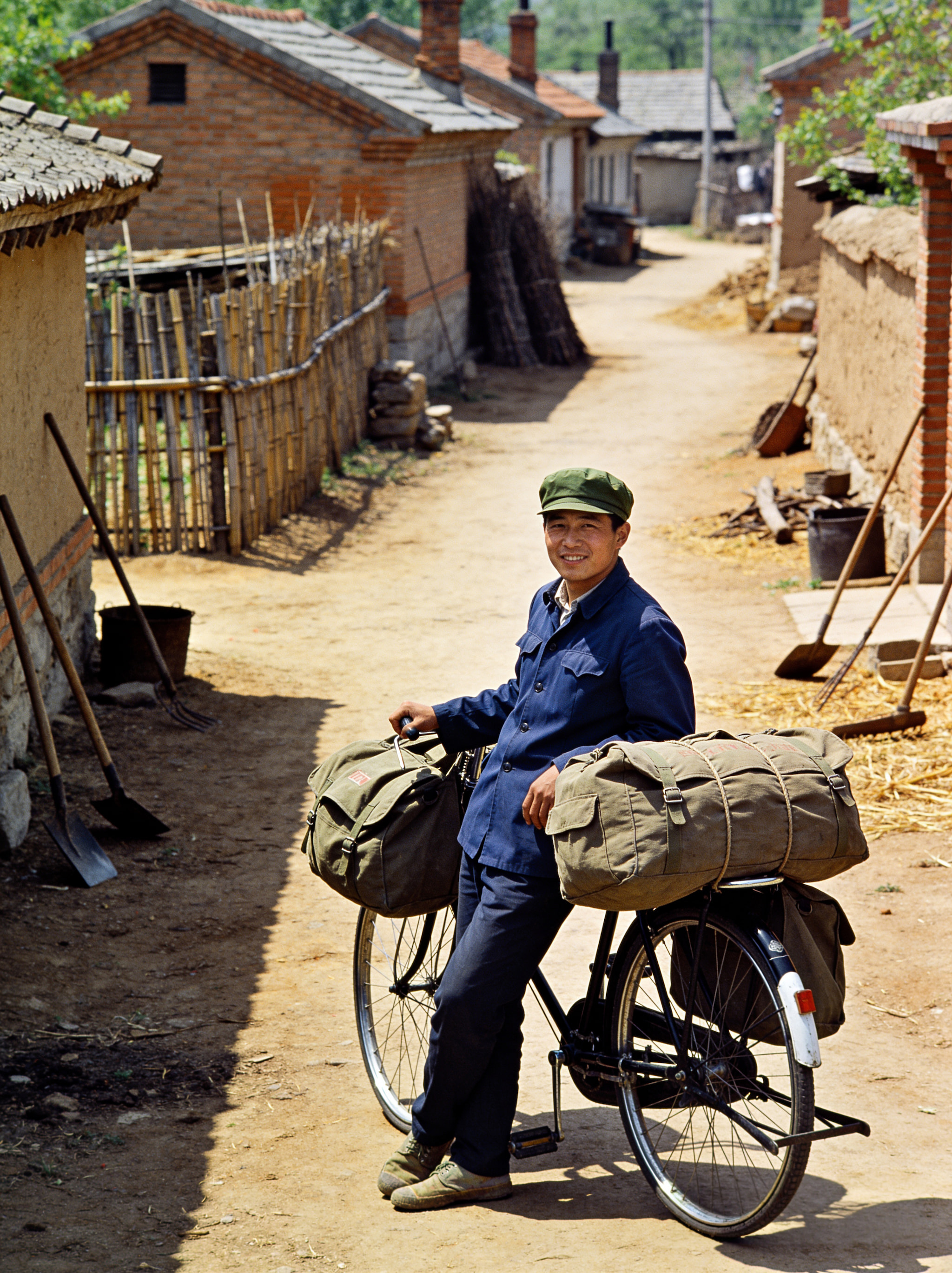 A man in a blue jacket and green cap posing with a bicycle loaded with canvas bags in a rural village alley.