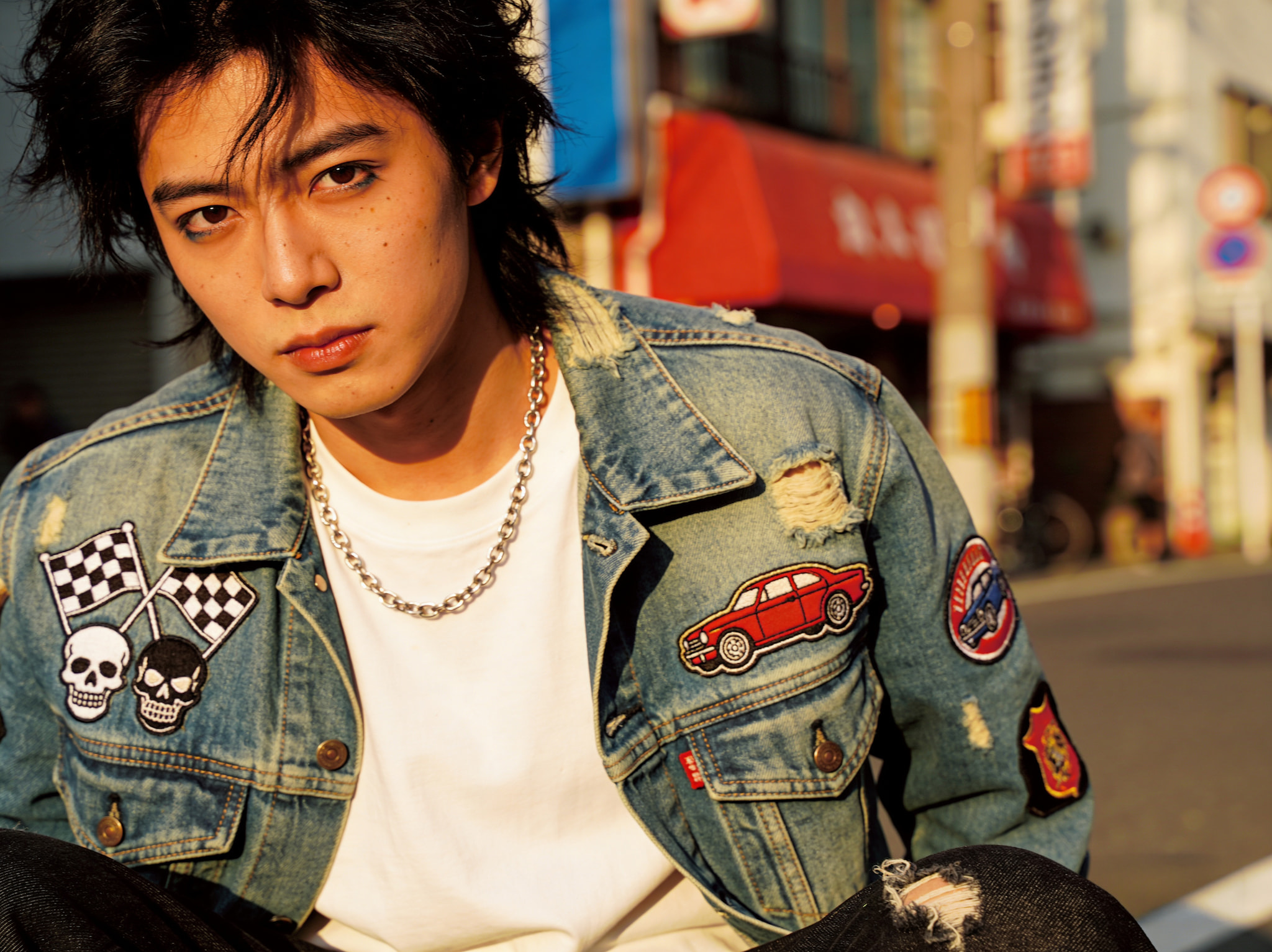 A close-up portrait of a young man with dark, messy hair wearing a distressed denim jacket with patches and a silver chain necklace in an urban street setting in Japan.