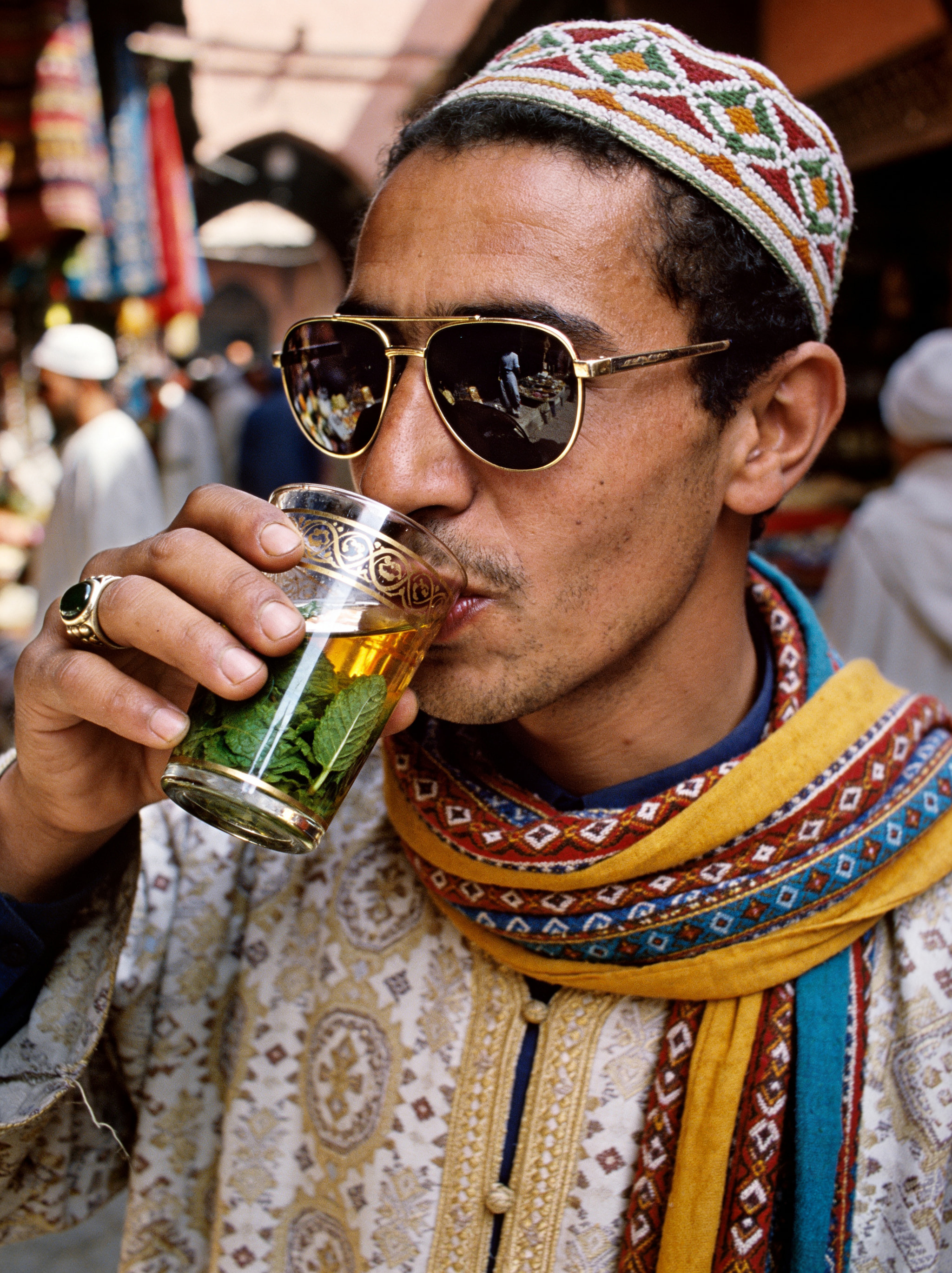 Man wearing traditional clothing and sunglasses drinking mint tea from an ornate glass in a market.