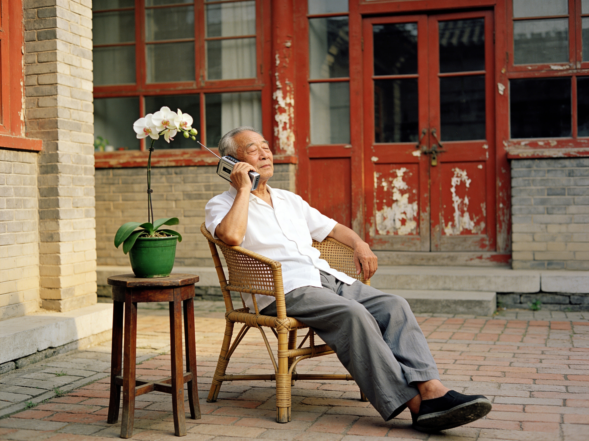 An elderly Chinese man relaxing in a wicker chair and holding a vintage transistor radio to his ear in a courtyard.