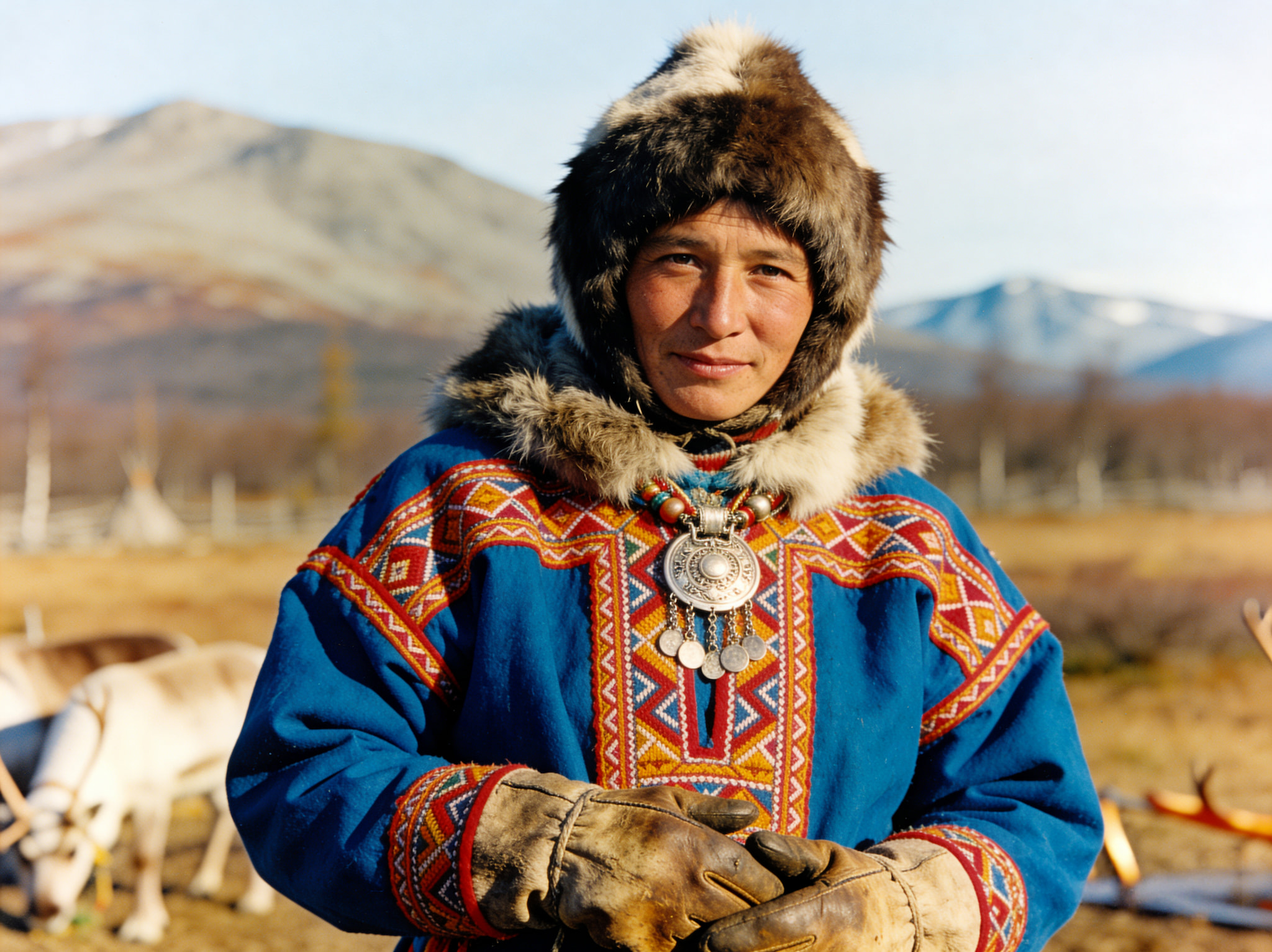 Portrait of a Sami person in traditional blue clothing with a fur-trimmed hood and silver necklace against a mountain landscape.