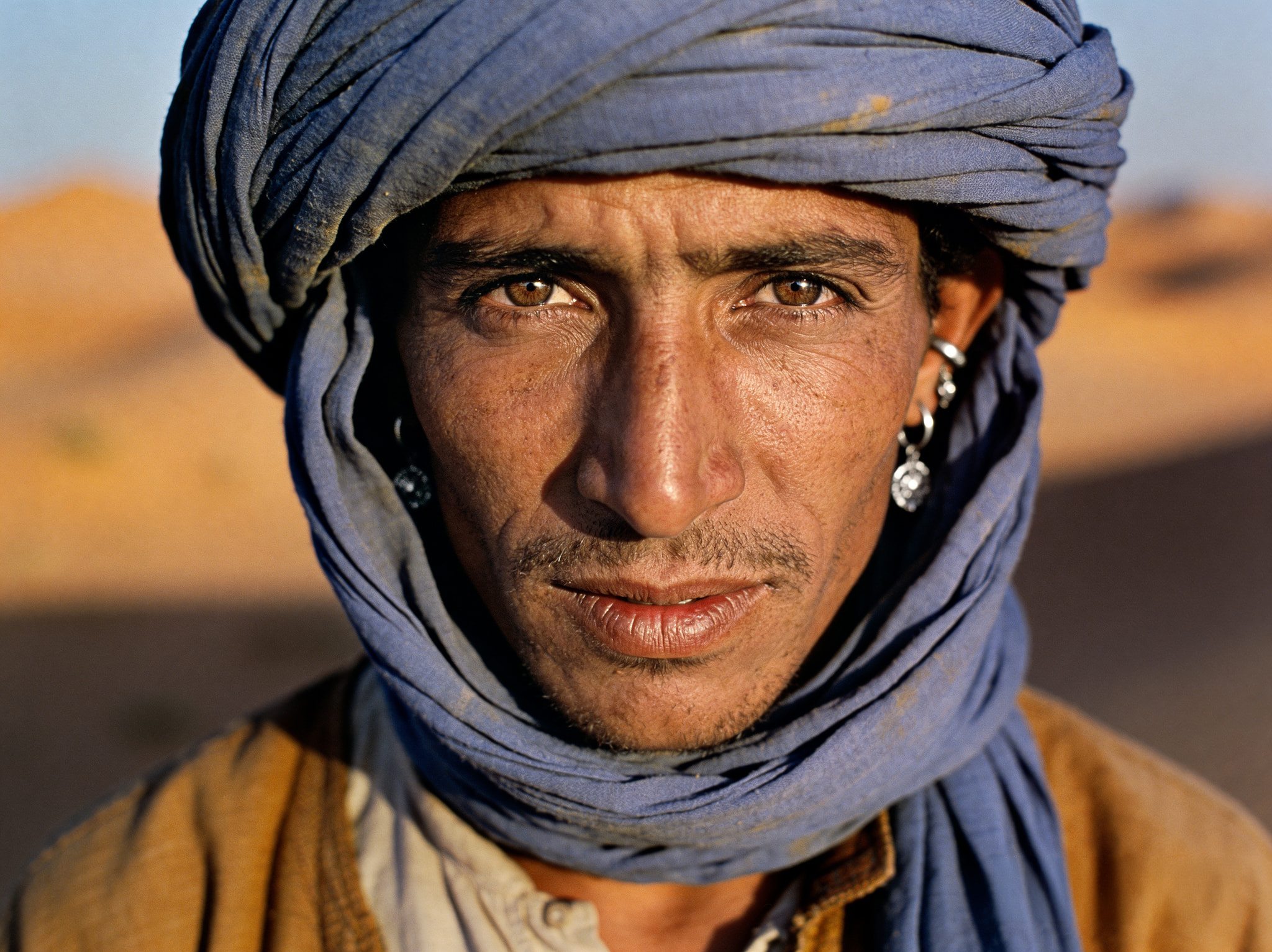 Portrait of a man with light brown eyes wearing a blue turban in a desert environment
