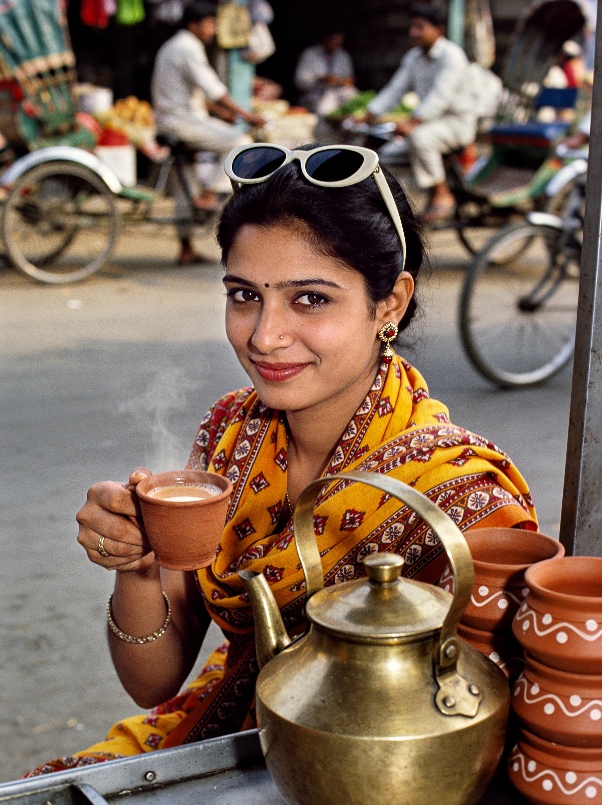 A smiling woman in a yellow traditional outfit holding a steaming cup of tea at a street stall.